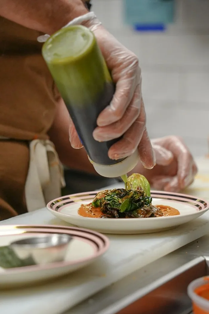 A cook in gloves piping green sauce onto a plate of greens