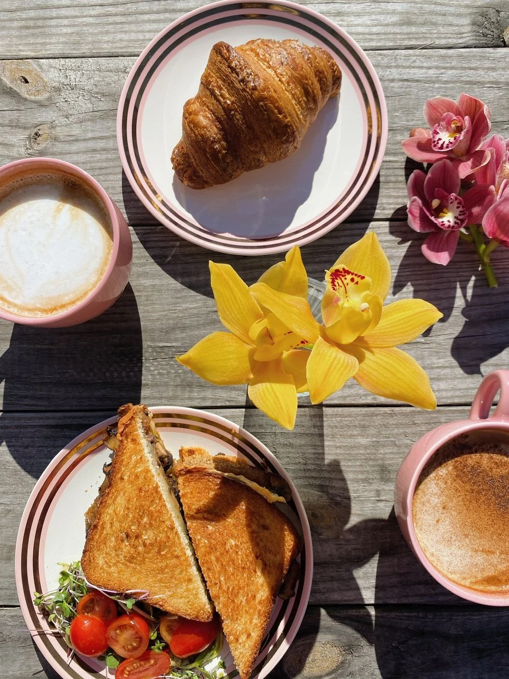 A croissant, a grilled sandwich and two lattes on striped plates with yellow and pink orchids, from above