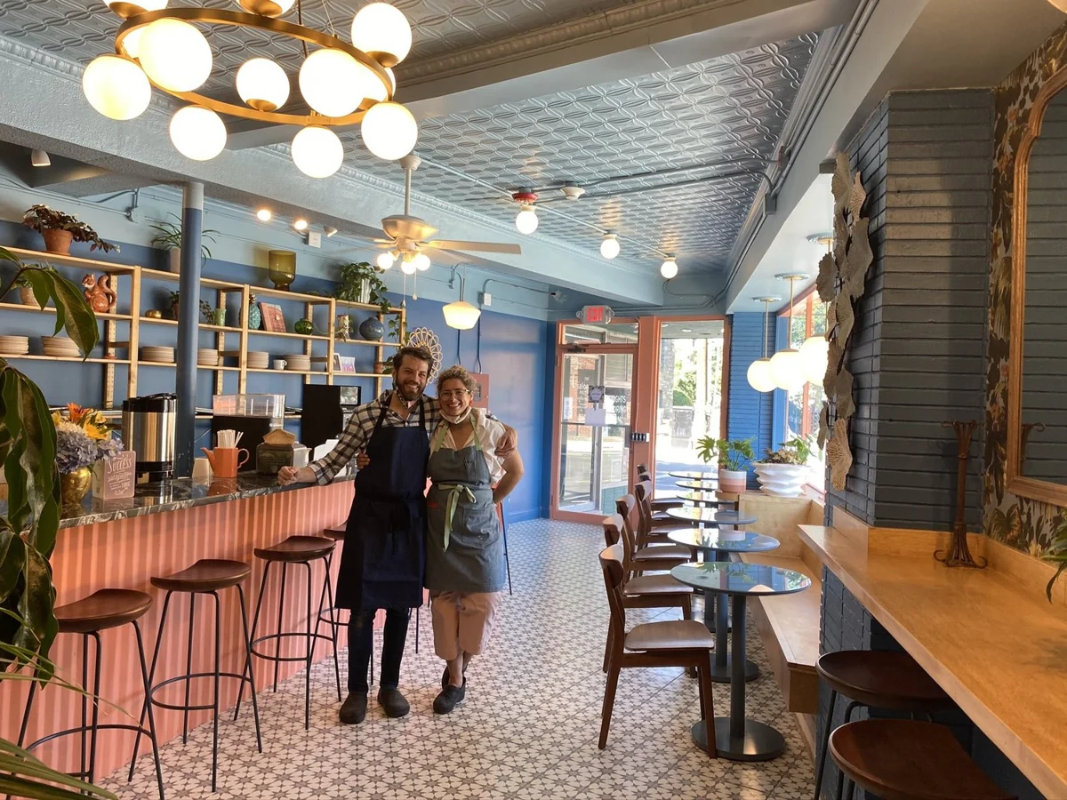 Two cooks in aprons, arm in arm in front of the pink counter and blue shelves of the café