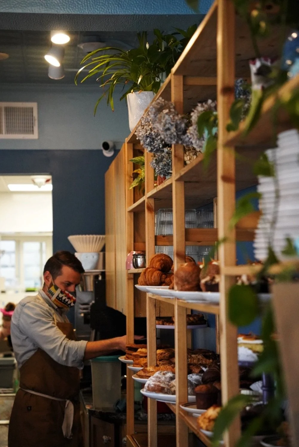 A barista at the coffee station beside open wooden shelves of croissants and pastries
